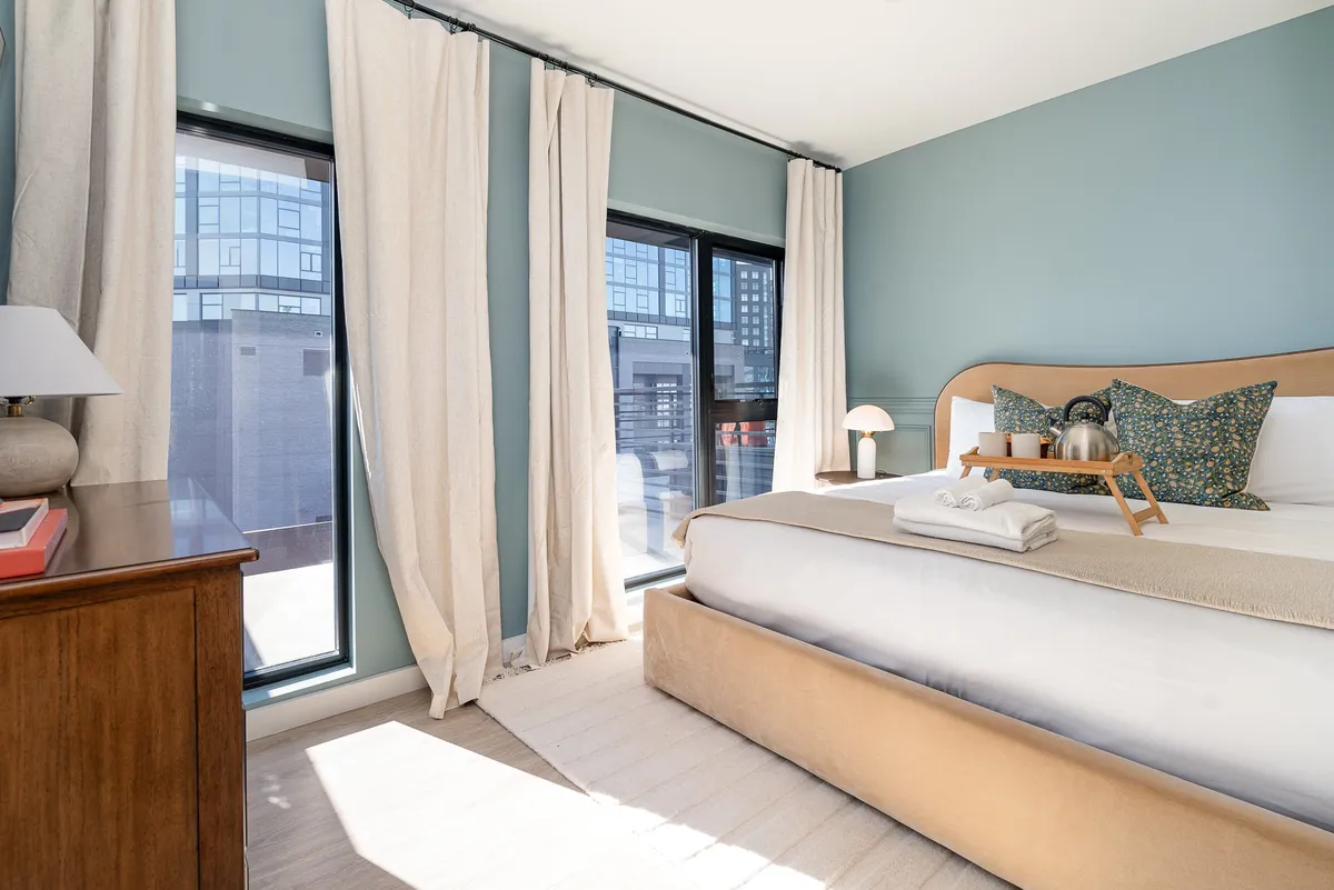 A serene bedroom featuring a large bed with crisp linens, ample natural light streaming in through generous windows, and a nightstand with a lamp. The minimalist decor and uncluttered design create a peaceful retreat, ideal for relaxation and privacy.
