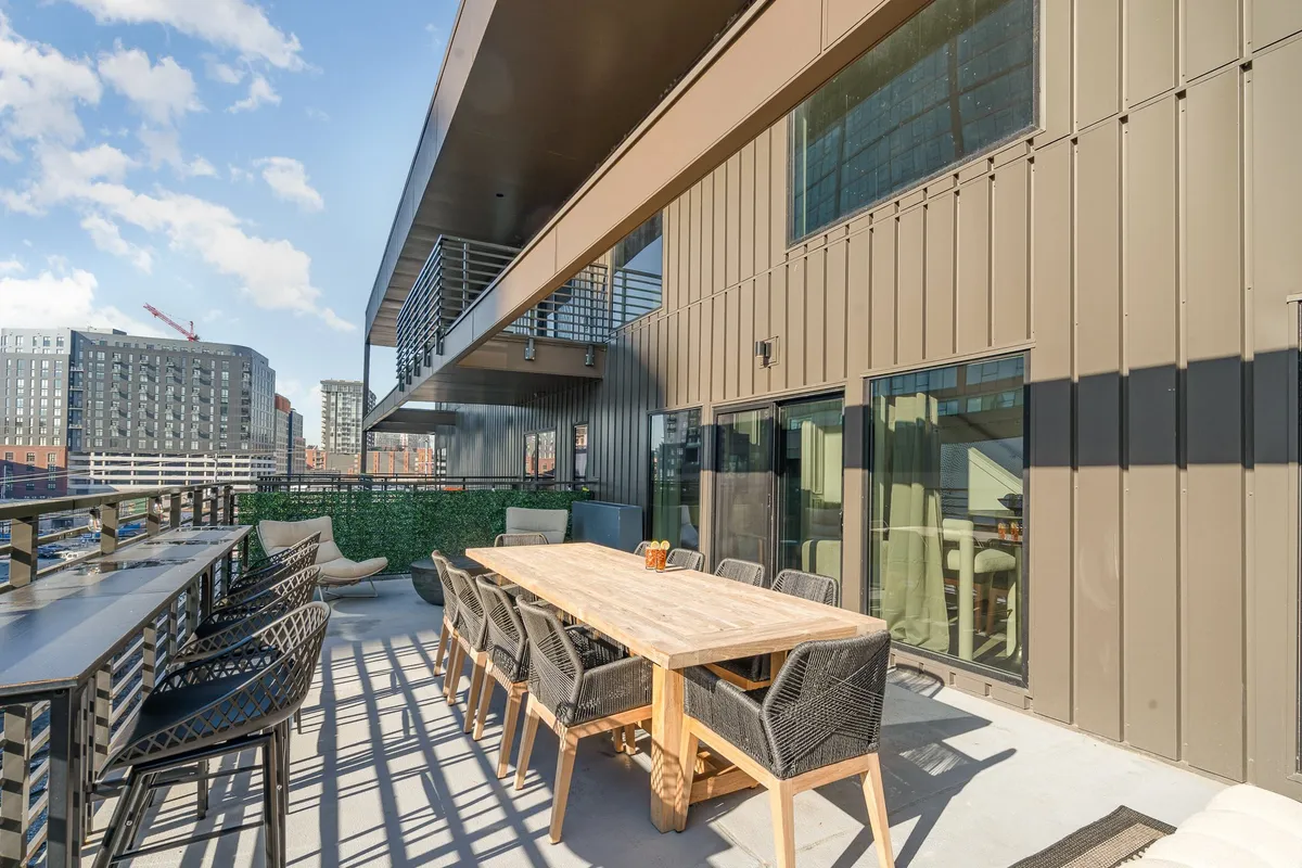 A spacious balcony featuring a large wooden dining table with black wicker chairs, lush green plants, and a stunning city skyline backdrop. The setup accommodates a group for outdoor dining and casual gatherings.
