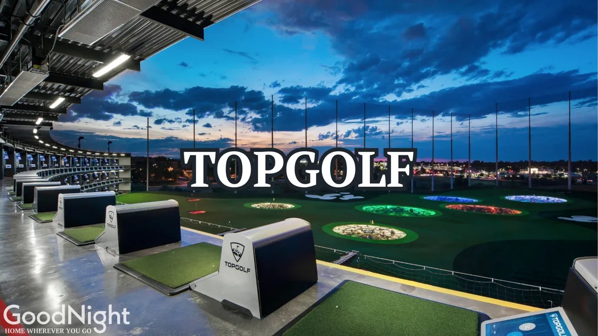 An exterior view of Topgolf’s driving range, with multiple golf hitting bays, glowing lights illuminating the range, and a dramatic sky with clouds overhead. Groups of people are seen enjoying golfing and socializing in an energetic setting.