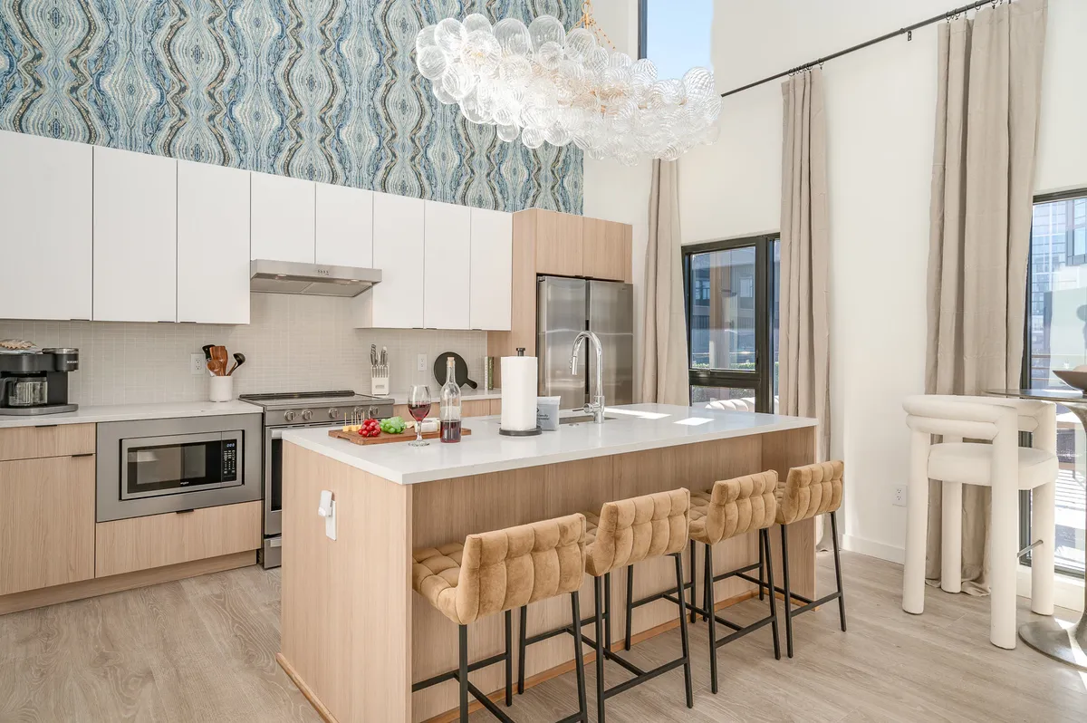 A contemporary kitchen with a sleek island, bar stools for four, a striking chandelier, and patterned wallpaper. The kitchen is open to the living area, perfect for casual dining or entertaining guests.