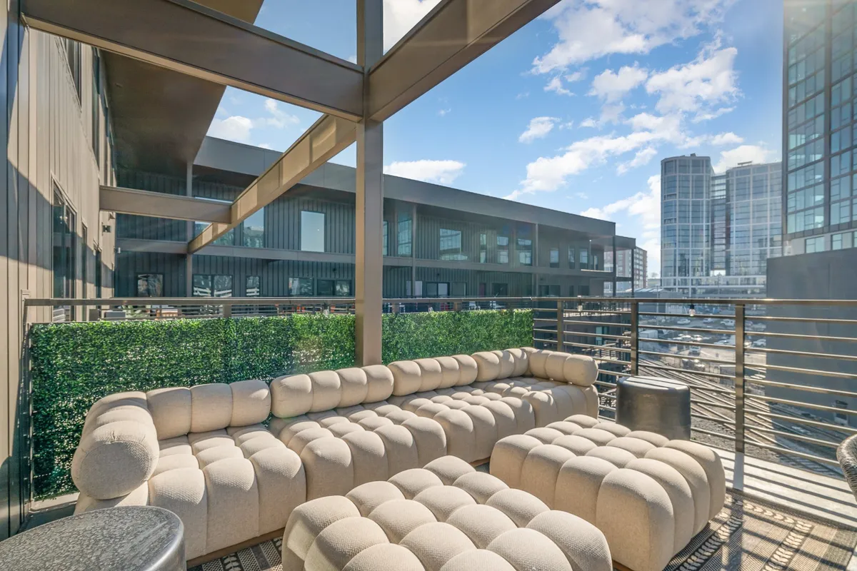 A contemporary outdoor seating area on a balcony or rooftop, featuring a glass railing with sweeping views of the city skyline. The space suggests relaxation and socializing after a day exploring the city, echoing the return-home narrative of the section.