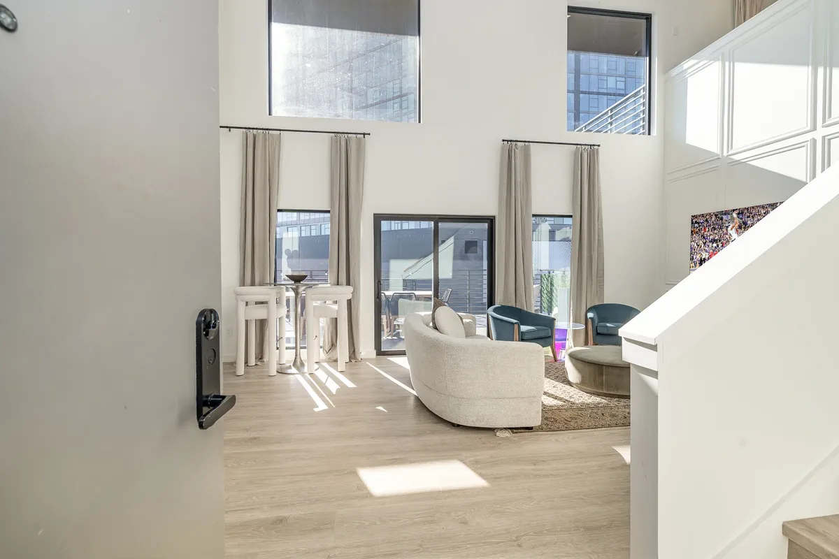 A cozy and modern living area with a sofa, barstools at the kitchen counter, large windows, and a staircase. The space looks inviting and functional, designed for both relaxation and social interaction.