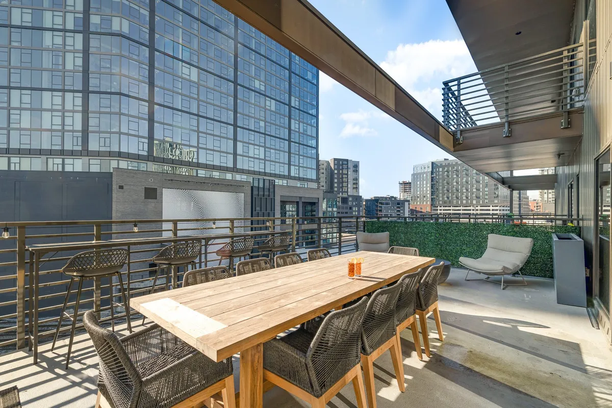 A spacious balcony with a large wooden dining table, modern chairs, lush greenery, and sweeping views of the Nashville city skyline. The setting is perfect for outdoor dining, group gatherings, and enjoying drinks against the urban backdrop.