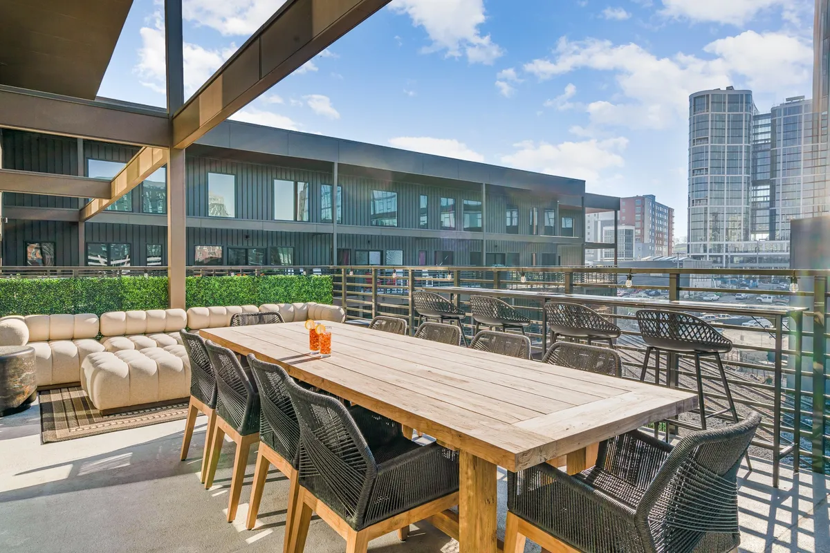 An expansive rooftop dining and lounge area with a large wooden table, comfortable seating, and a sweeping city skyline in the background. The space is ideal for sunset viewing, social gatherings, and outdoor meals.