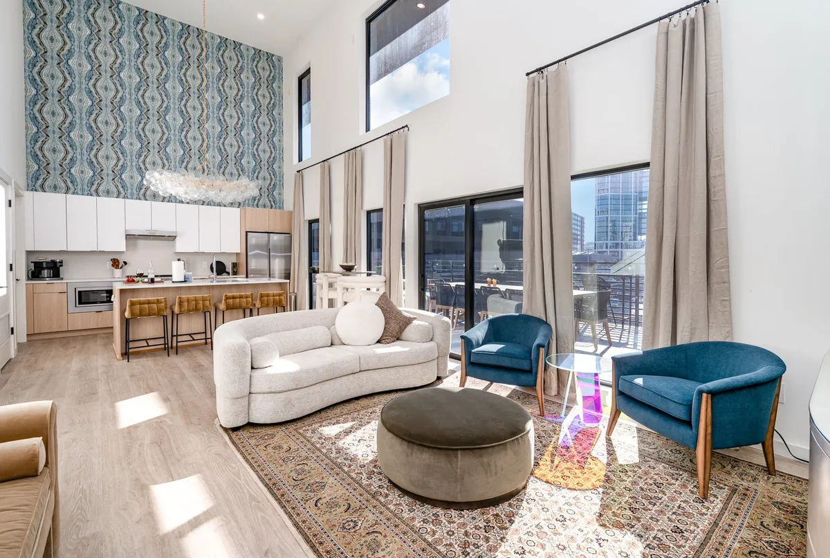 The image showcases a stylish, sunlit living area featuring a tall patterned accent wall, plush bright sofa, colorful chairs, and a round ottoman that invite relaxation and socializing. In the background, a sleek kitchen bar area hints at shared meals or lively brunches, all set within a contemporary, welcoming atmosphere perfect for gathering with friends or family.