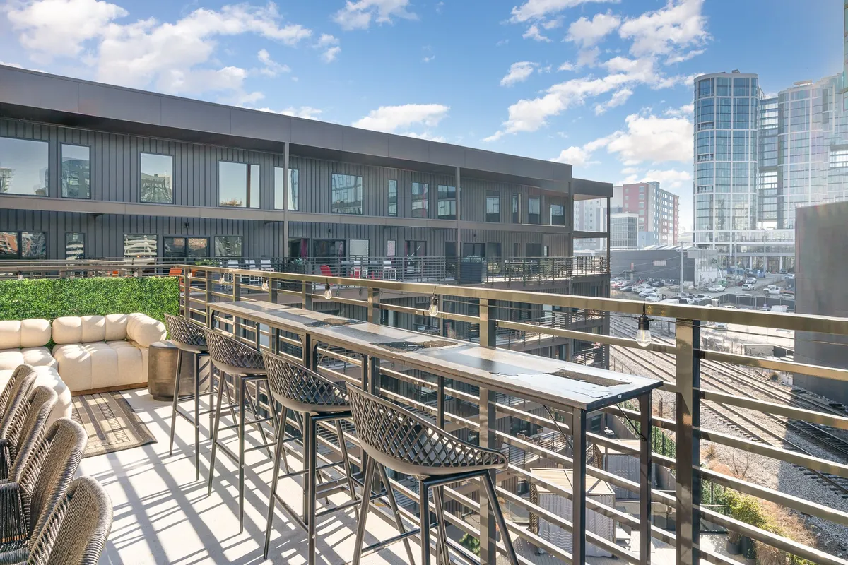 A stylish rooftop bar area with comfortable seating overlooks the Nashville city skyline. The atmosphere is relaxed yet social, with plenty of space for gatherings and sweeping sunset views. The modern furniture and bar setup hint at the perfect spot for unwinding after a productive day, while the urban backdrop emphasizes downtown convenience.