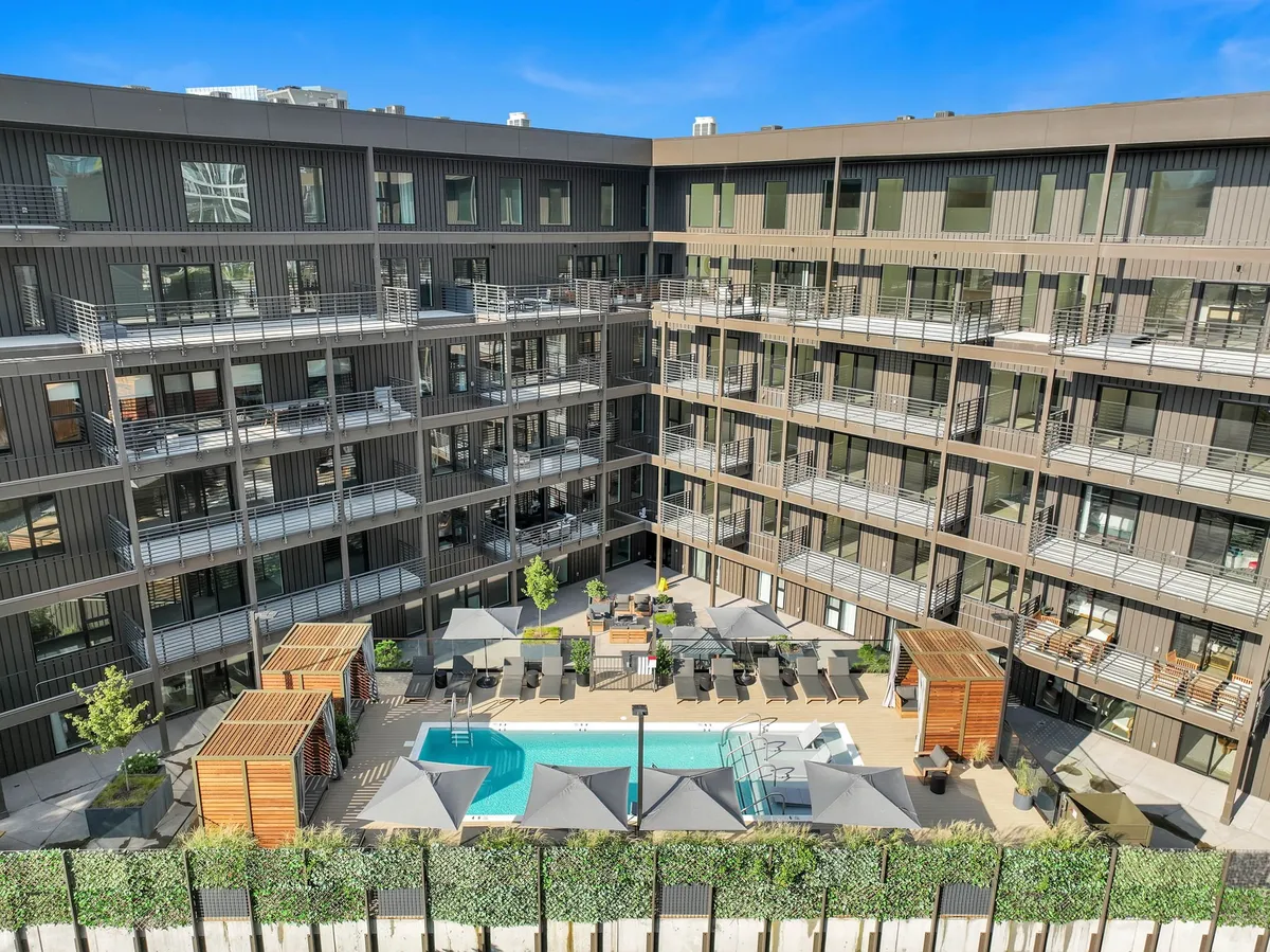 A vibrant outdoor swimming pool area featuring lounge chairs, landscaped areas, and stylish wooden cabanas, set within a lively building environment. The scene suggests an inviting space for pool parties, sunbathing, or relaxing by the water with friends.